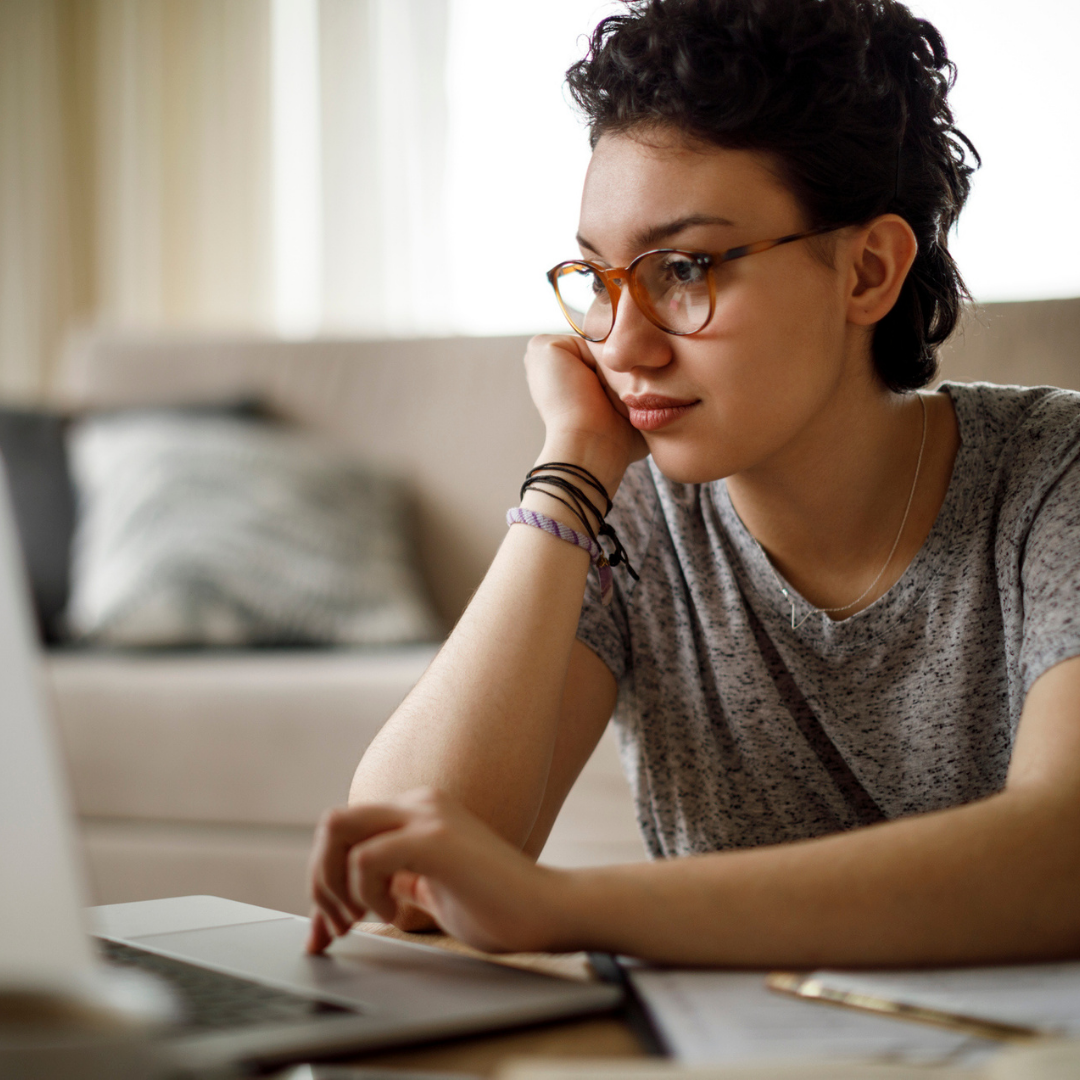Promo image of a young person with short wavy brown hair, brown eyes, and glasses, working at a laptop with a thoughtful expression.