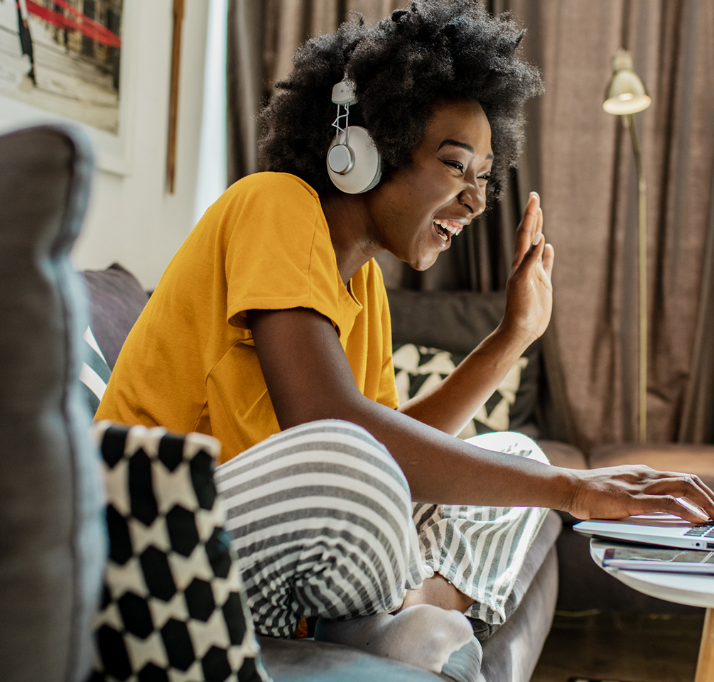 Promo image of a young Black woman wearing silver headphones, a yellow shirt and gray and white striped pants, smiling and waving to her computer screen while sitting on a gray couch.